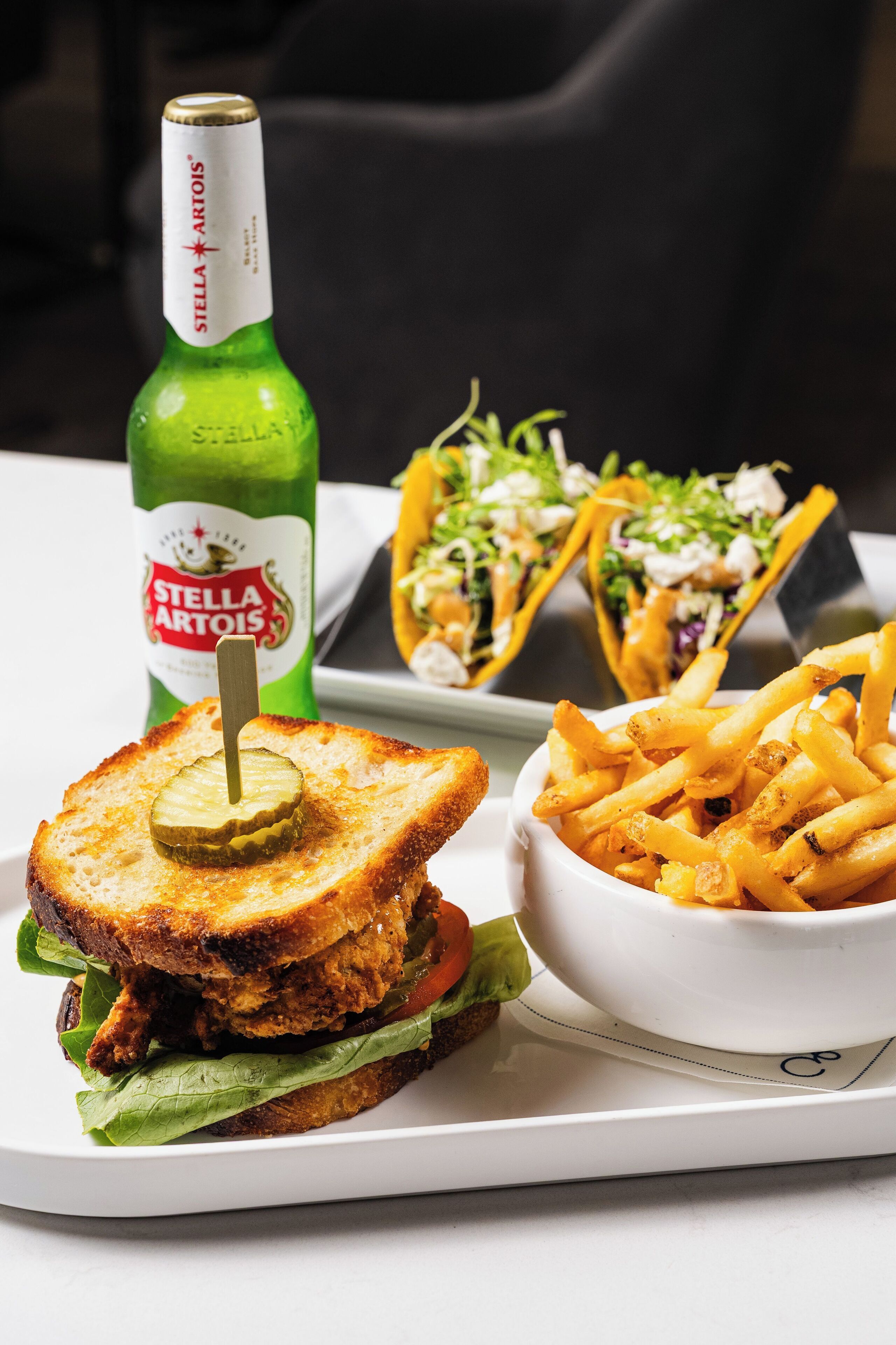 Un sandwich avec des cornichons, une bouteille de bière Stella Artois, un bol de frites et une salade-taco, le tout servi sur un plateau.