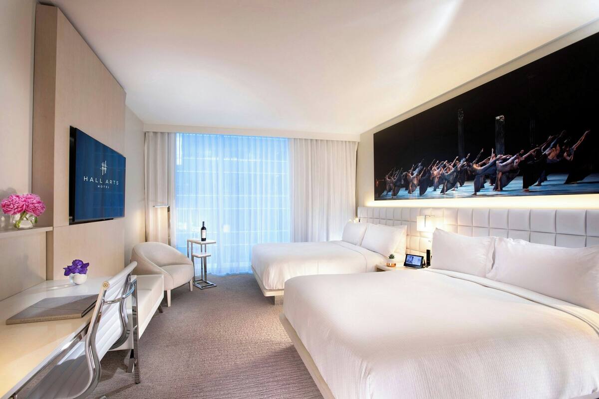 A hotel room with two beds, a desk, a chair, a TV, and a large window with curtains.