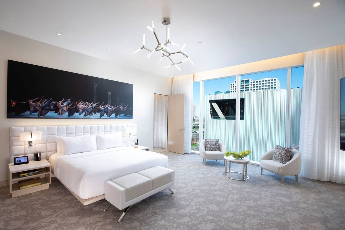A modern hotel room with a large bed, a desk, a chair, and a large window with curtains.