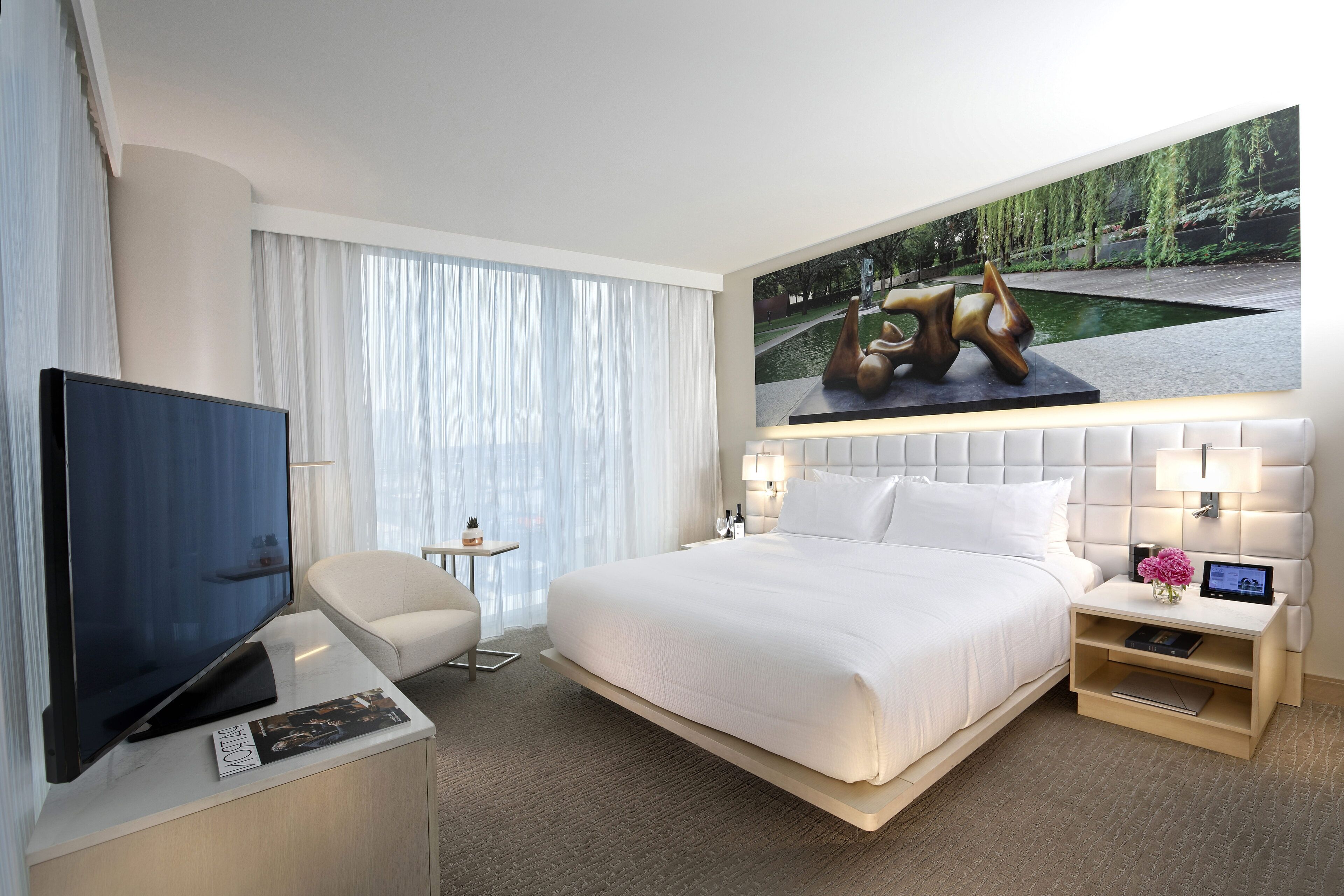 A modern hotel room with a large bed, a flat-screen TV, a small desk, and a nightstand with a lamp and a flower vase.