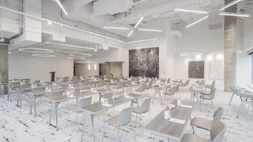 A spacious conference room with multiple tables and chairs, modern lighting fixtures, and a large abstract painting on the wall.
