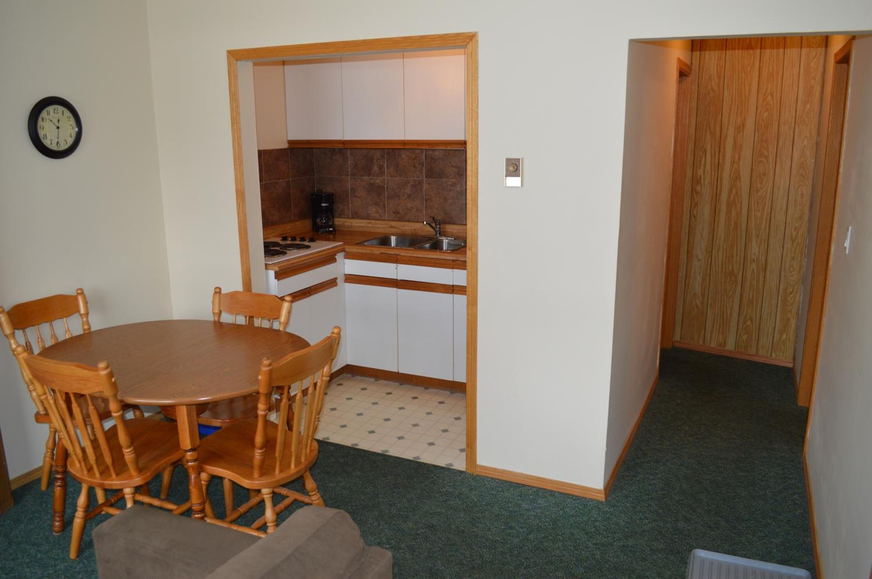 Una cocina compacta con armarios blancos, una mesa de comedor de madera con sillas y un reloj en la pared.