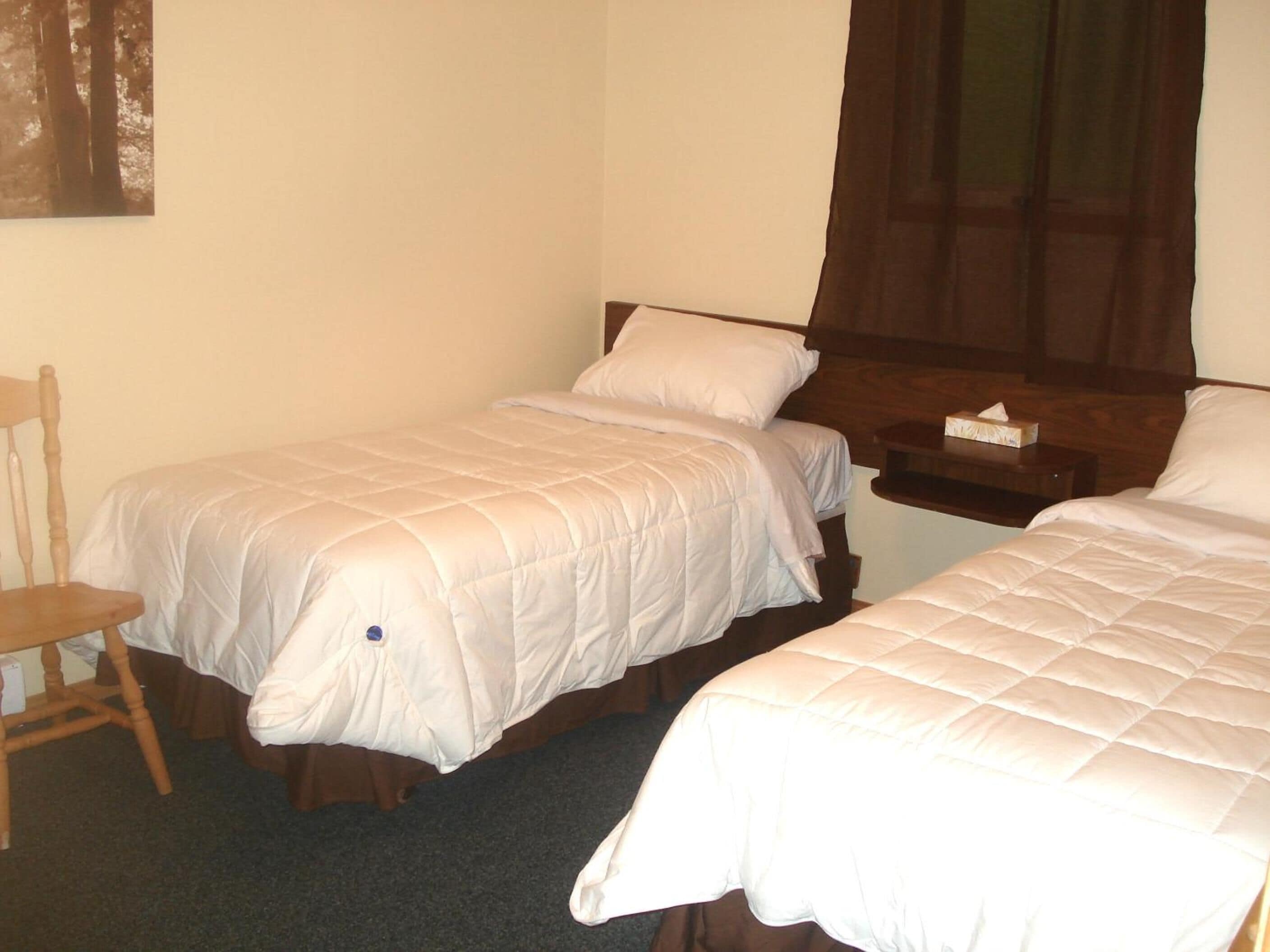 Una habitación con dos camas individuales, una silla de madera, una mesita de noche con un dispensador de pañuelos y una ventana con cortinas.