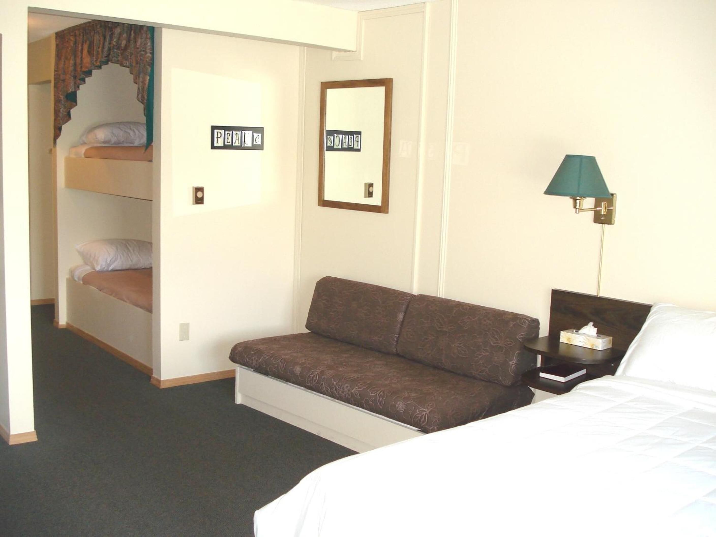 A hotel room with a bunk bed, a sofa, a bedside table with a lamp, and a framed picture on the wall.
