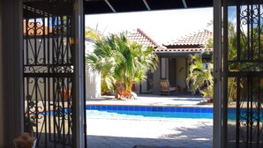 Una puerta abierta que da a una zona de piscina con suelo de baldosas, palmeras y un edificio con tejado de cerámica al fondo.