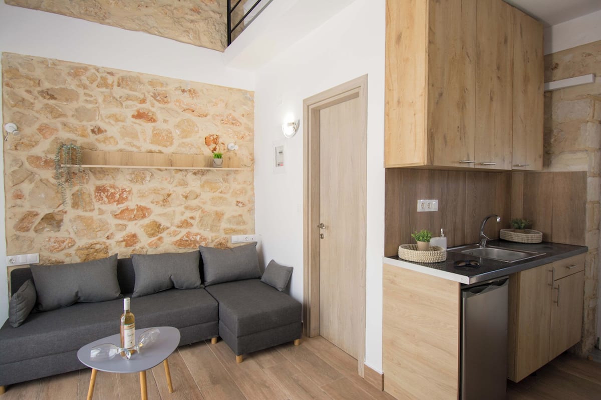 A compact living space with a stone wall, grey sofa, wooden cabinets, and a small kitchen area.