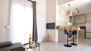 A modern living room with a grey sofa, a small round table with a bottle and glasses, and a TV mounted on the wall. - Michalianna suites (Hersonissos)