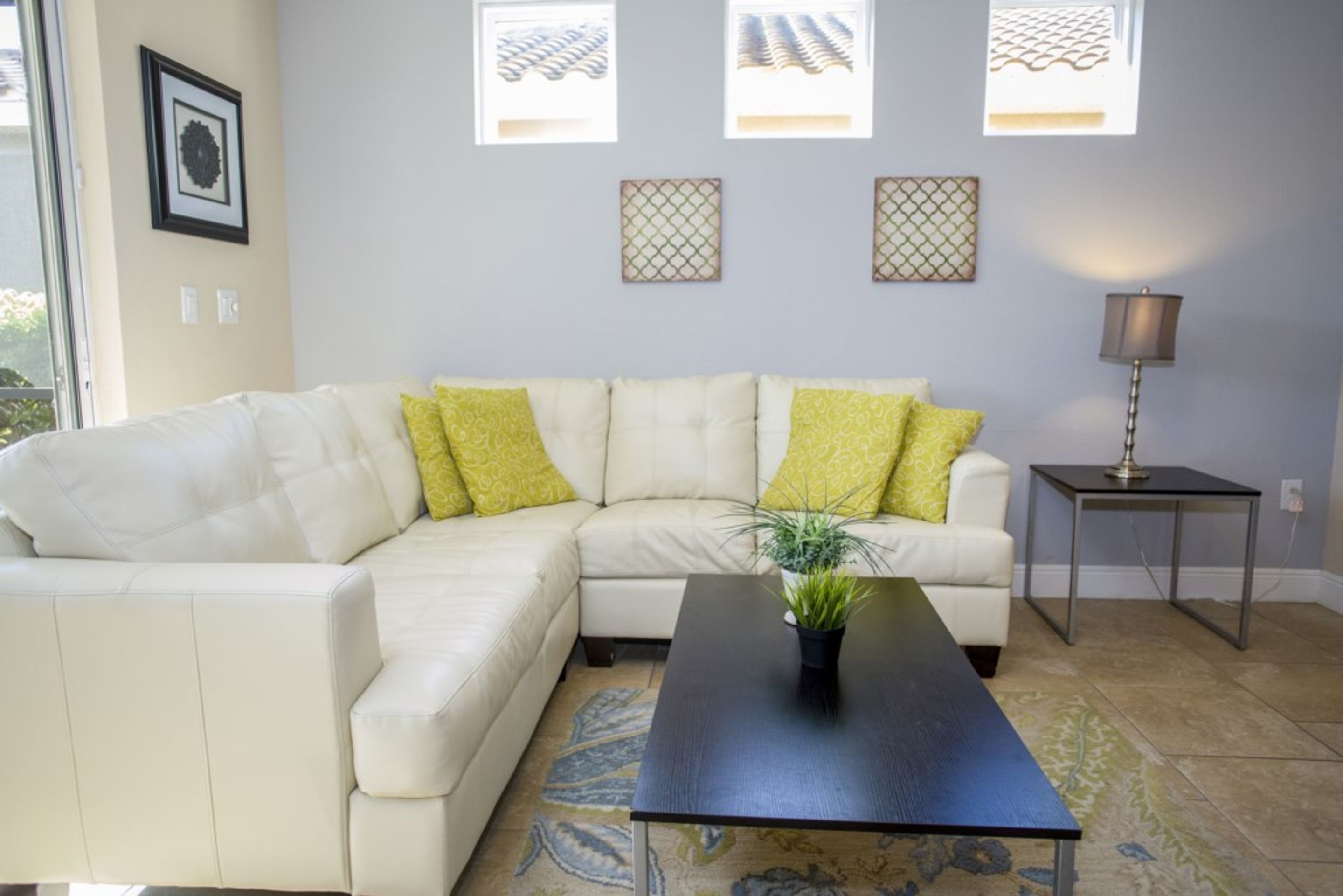 A living room with a white sectional sofa, a black coffee table, and a small side table with a lamp. There are three framed pictures on the wall and a potted plant on the coffee table.