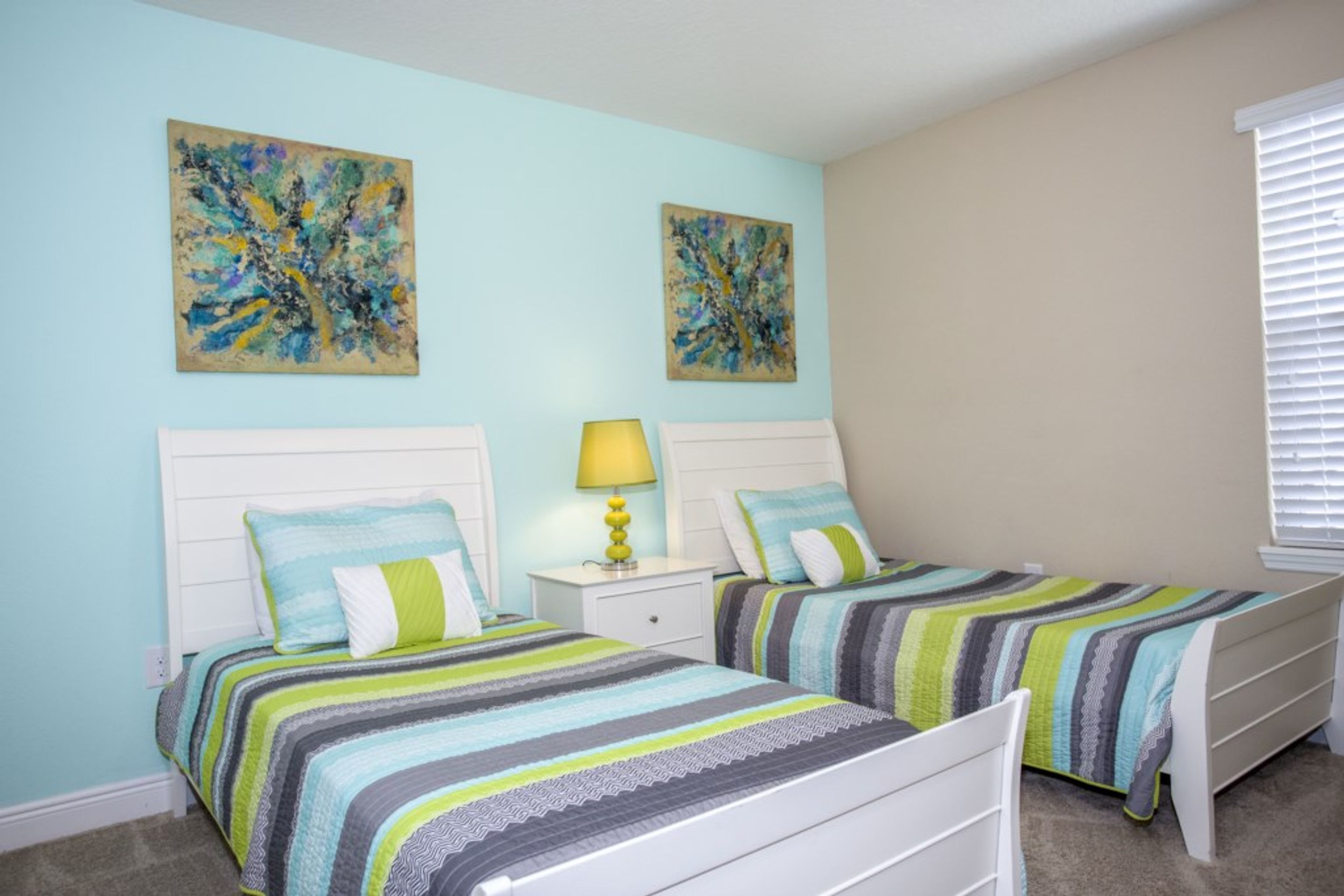 A bedroom with two beds, a nightstand with a lamp, and two wall-mounted paintings.