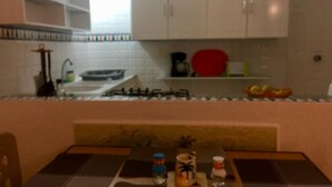 Une cuisine avec des meubles blancs, une cuisinière à gaz et un plan de travail sur lequel on trouve une assiette rouge, une machine à café et quelques fruits.