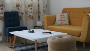 Un salon avec un canapé, un fauteuil bleu, une petite table recouverte d’une nappe blanche et une paire d’écouteurs posés sur la table.