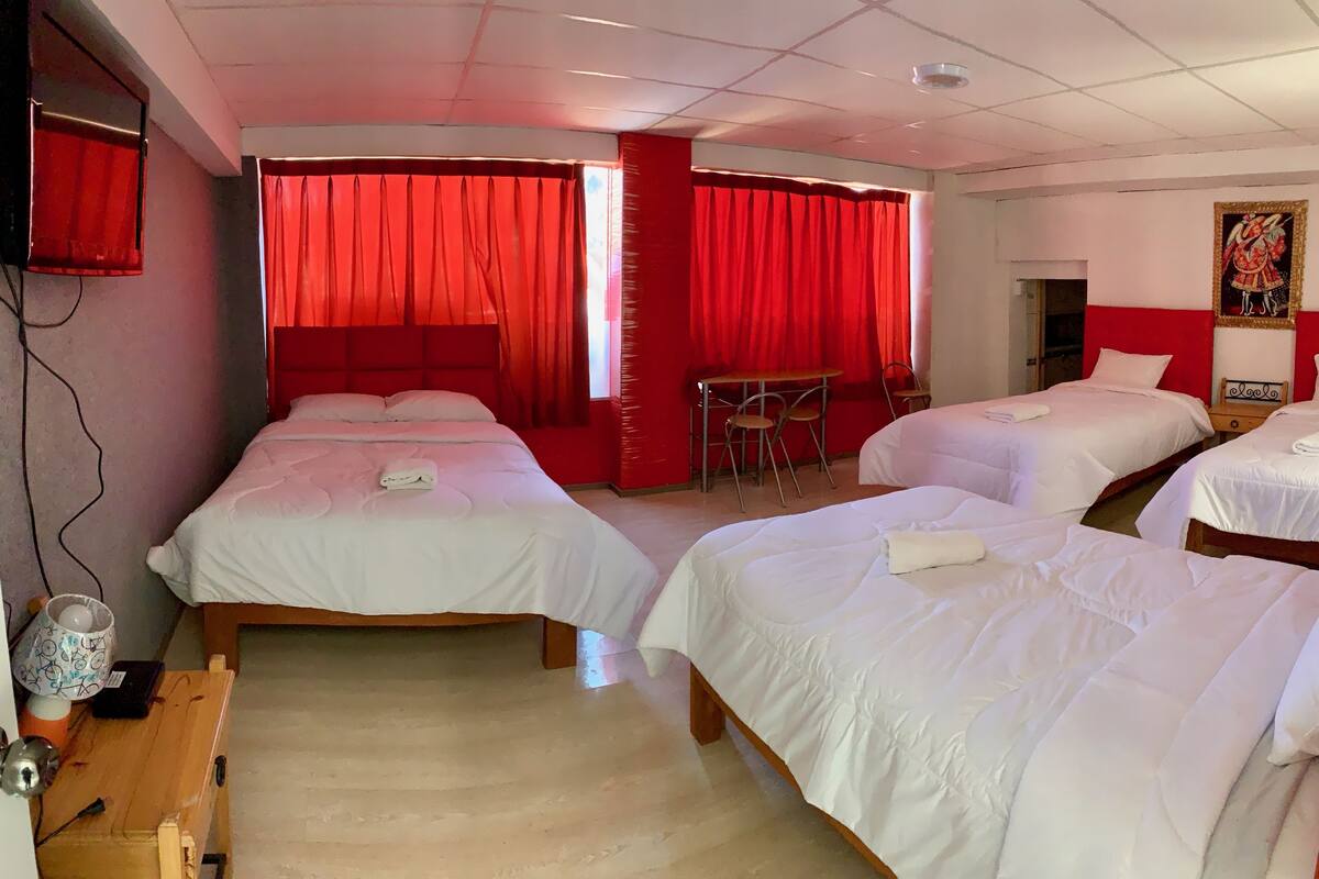 Une chambre d’hôtel avec trois lits simples, une petite table avec des chaises et un rideau rouge.