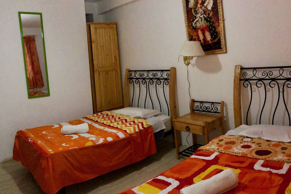 Une chambre avec deux lits simples, chacun recouvert d’une couvre-lit à motifs et agrémenté d’une serviette roulée. On y trouve une armoire en bois et une petite table de chevet avec une lampe.