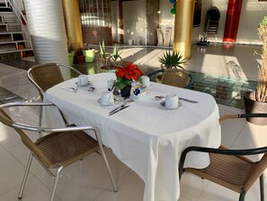 A round table set for a meal with white tablecloth, surrounded by wicker chairs, in a sunlit outdoor area with potted plants and a brick wall in the background. - Hotel Plateros (Cusco)