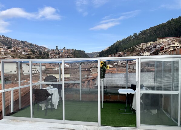 A glass-enclosed balcony with a view of a hillside town, a table, and a chair. - Hotel Plateros (Cusco)