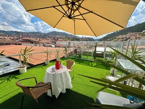 Een terras met een tafel, stoelen, een gele parasol en plantenpotten.