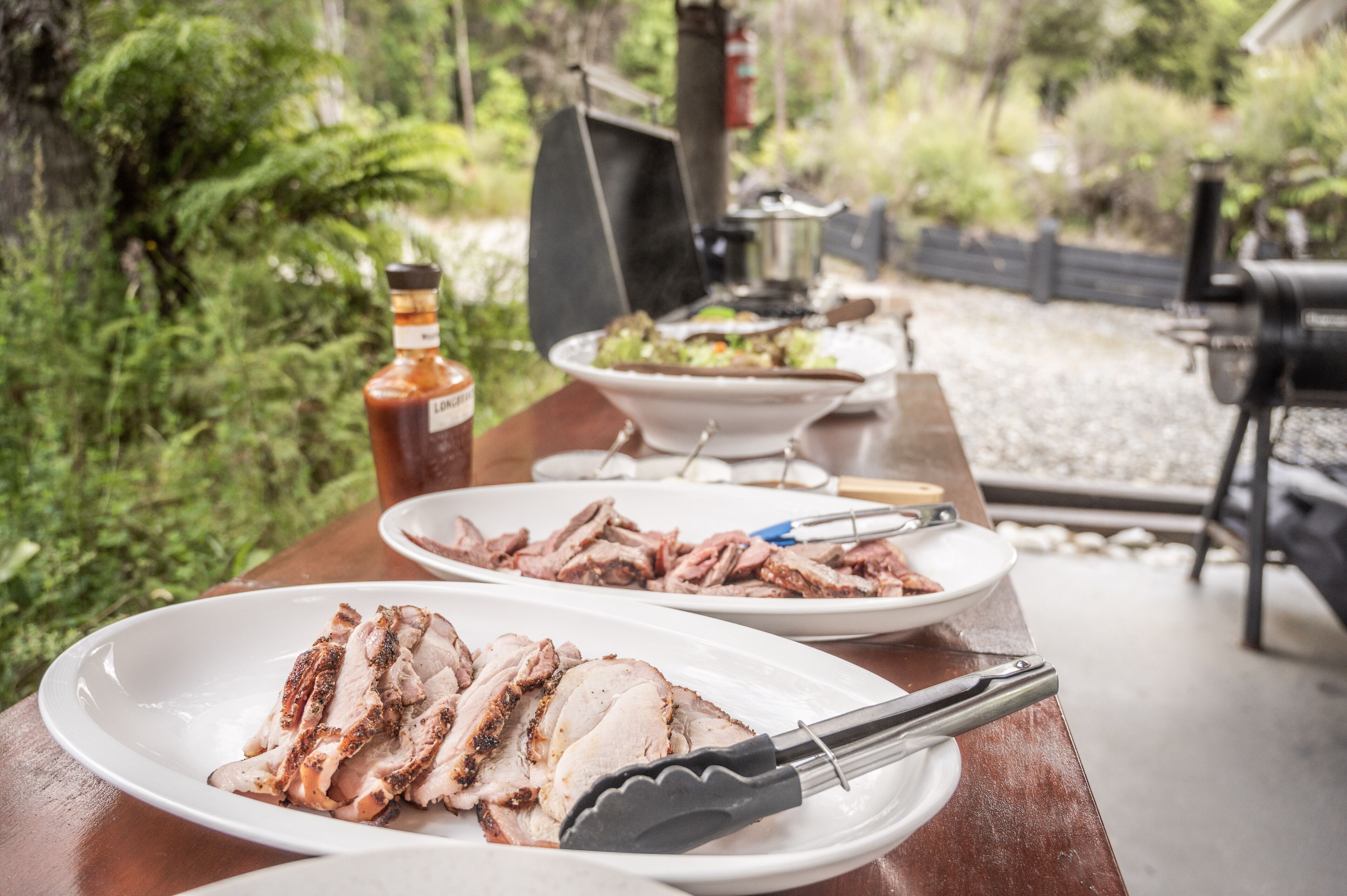 A table with sliced meat and a bottle of sauce, a bowl of salad, and a grill in a garden setting.