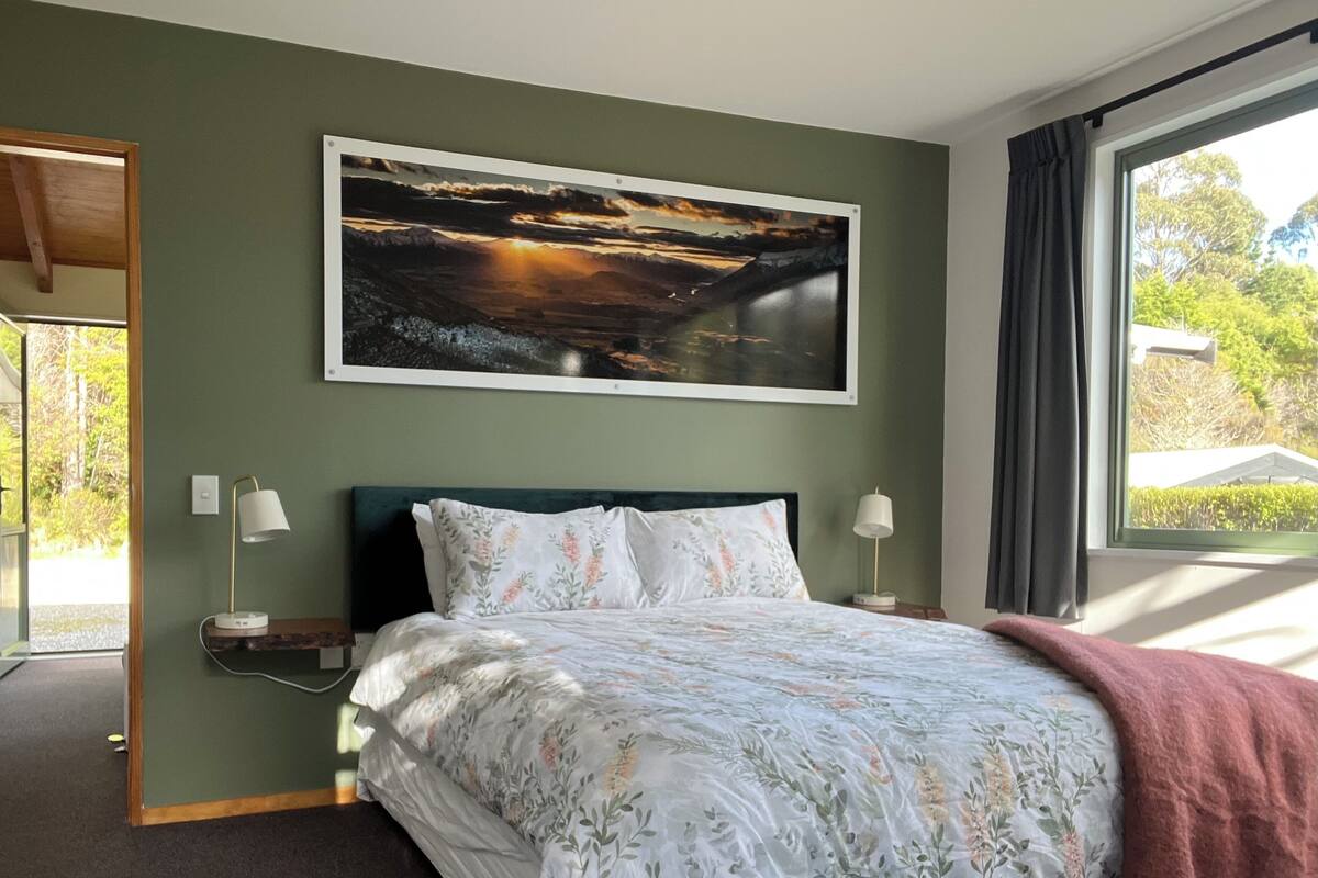 A bedroom with a bed, two bedside lamps, a nightstand, a painting above the bed, and a window with curtains.