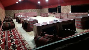A room with a high ceiling, red walls, and patterned floor. There are several tables and benches arranged for seating. - Atlantis Rum Camp (Wadi Rum)