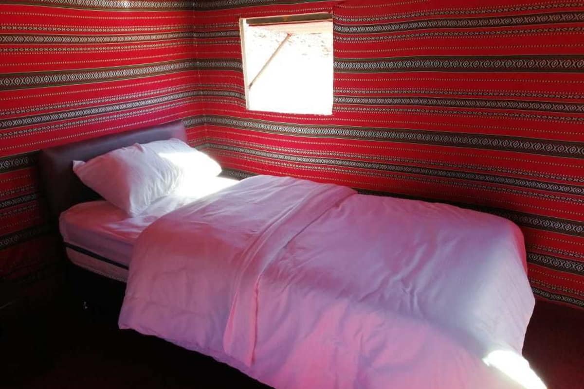 A single bed with a white mattress and pillows, set against a red patterned wall with a small window.