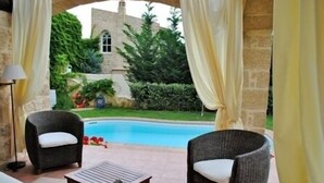 Un espace extérieur abrité comprenant deux fauteuils en osier et une petite table, donnant sur une piscine et un bâtiment en pierre avec un jardin.