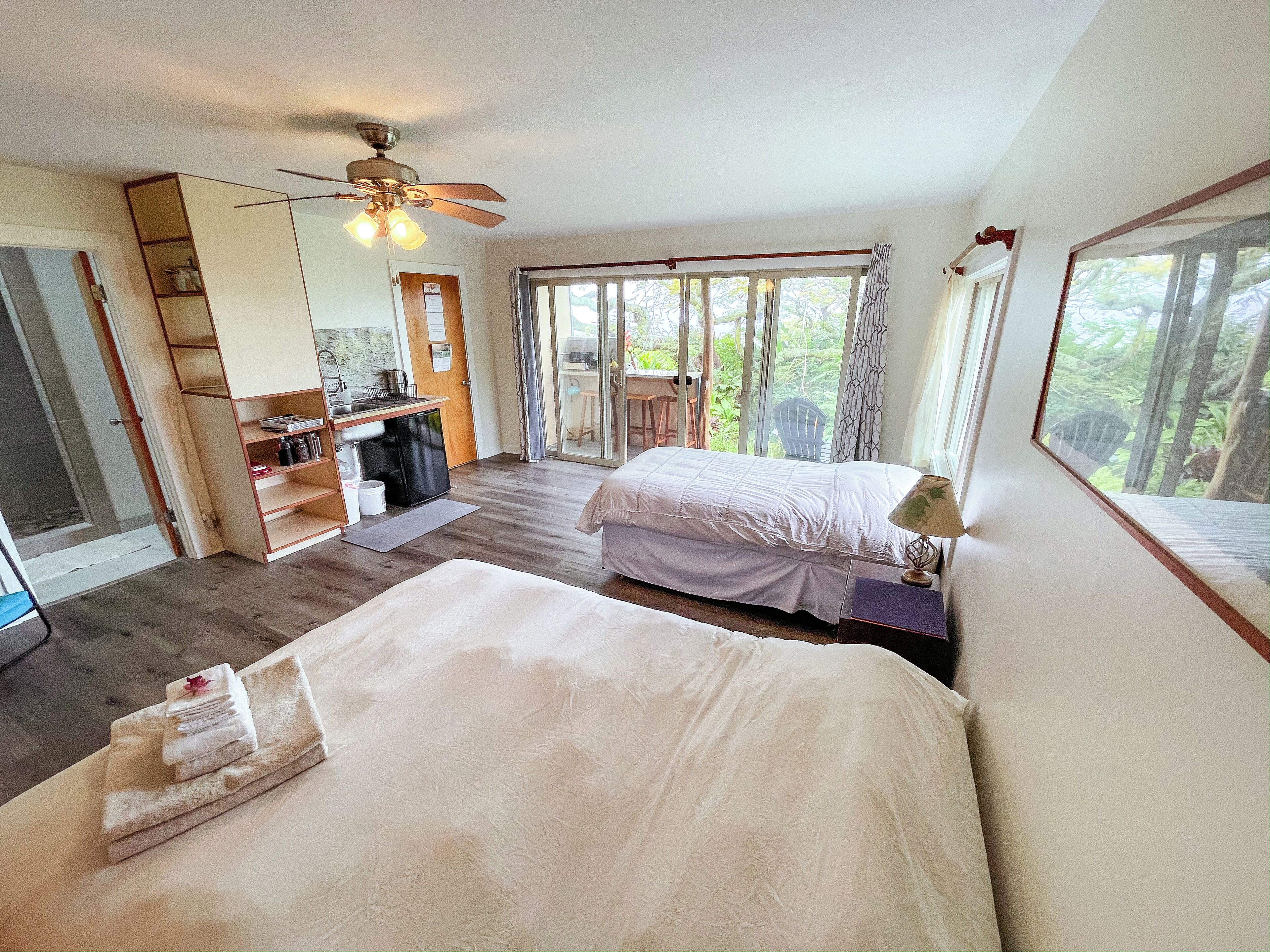 A bedroom with two beds, a ceiling fan, a small kitchenette, and a large window with a view of greenery.