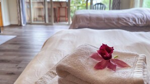 A neatly made bed with a folded towel and a rose, a wooden floor, and a view of an outdoor area with a chair and a table through a glass door. - Gingerhill Farm Retreat (Kealakekua)
