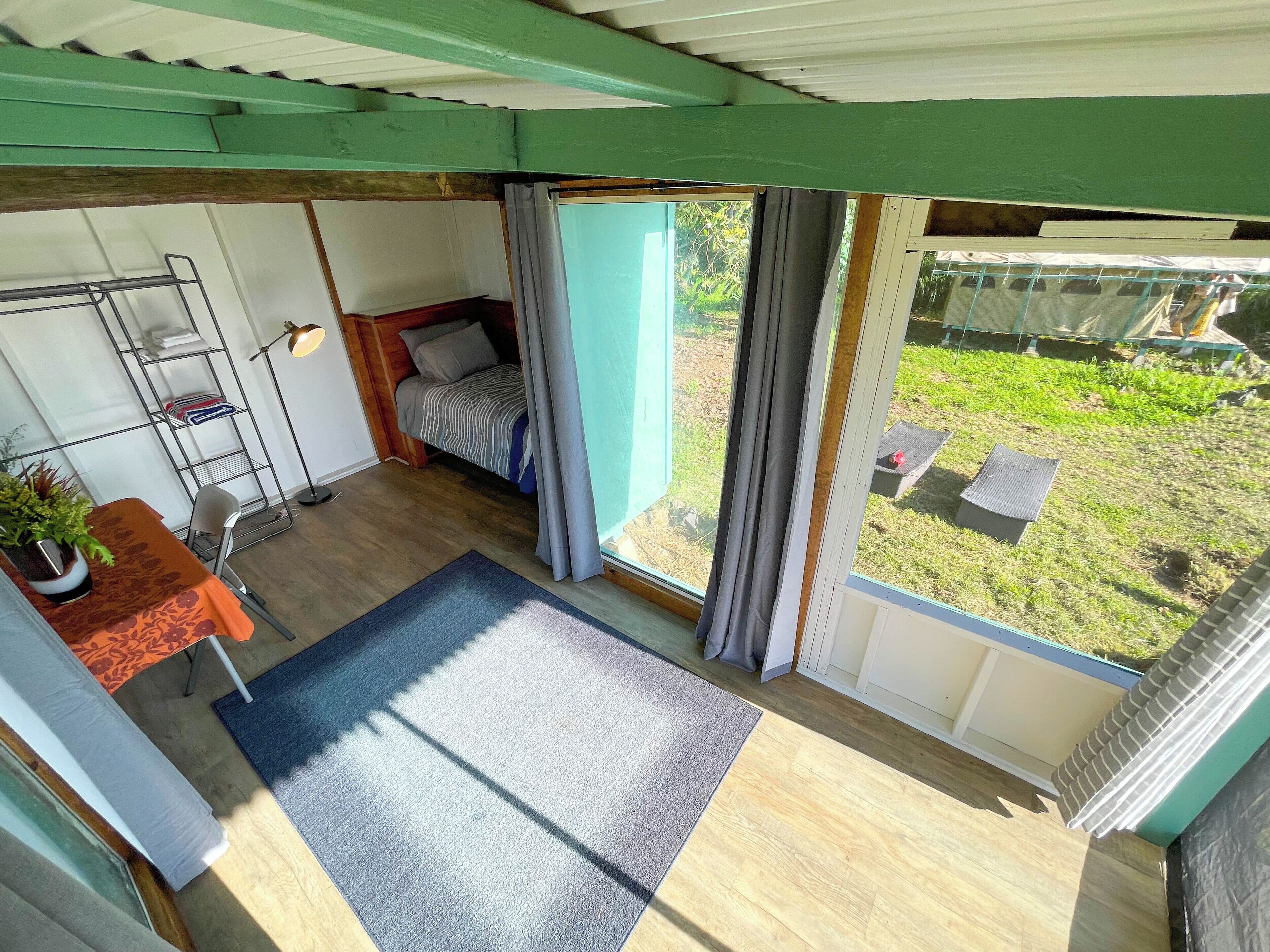 Une pièce comprenant un lit, une petite table avec une plante, une étagère en métal et une vue sur l’extérieur.