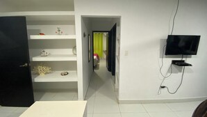 A room with a white door, a white table, a TV mounted on the wall, and a shelf with decorative items. - Jackuri Casa Hospedaje (San Andrés)