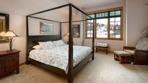Un lit à baldaquin avec une tête de lit sombre, une table de chevet en bois avec une lampe, une chaise avec un coussin à motifs et une fenêtre donnant sur un paysage enneigé.