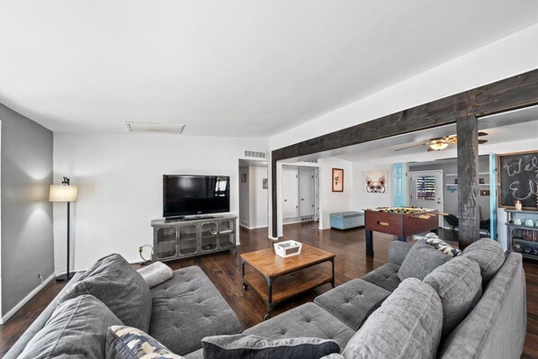 A modern living room with a large gray sectional sofa, a wooden coffee table, a flat-screen TV, and a foosball table. - Eli Drive: Farmhouse Charm Meets Modern Luxury – Pool Included! (Tucson)
