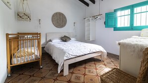 Una camera da letto con una culla, un letto, un decoro macramè appeso, un quadro a parete circolare, una finestra con persiane color acquamarina e piastrelle decorative.