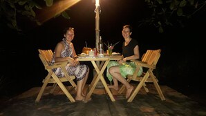 Deux personnes sont assises à une table en bois à l’extérieur, la nuit, et une lampe de table éclaire la scène.