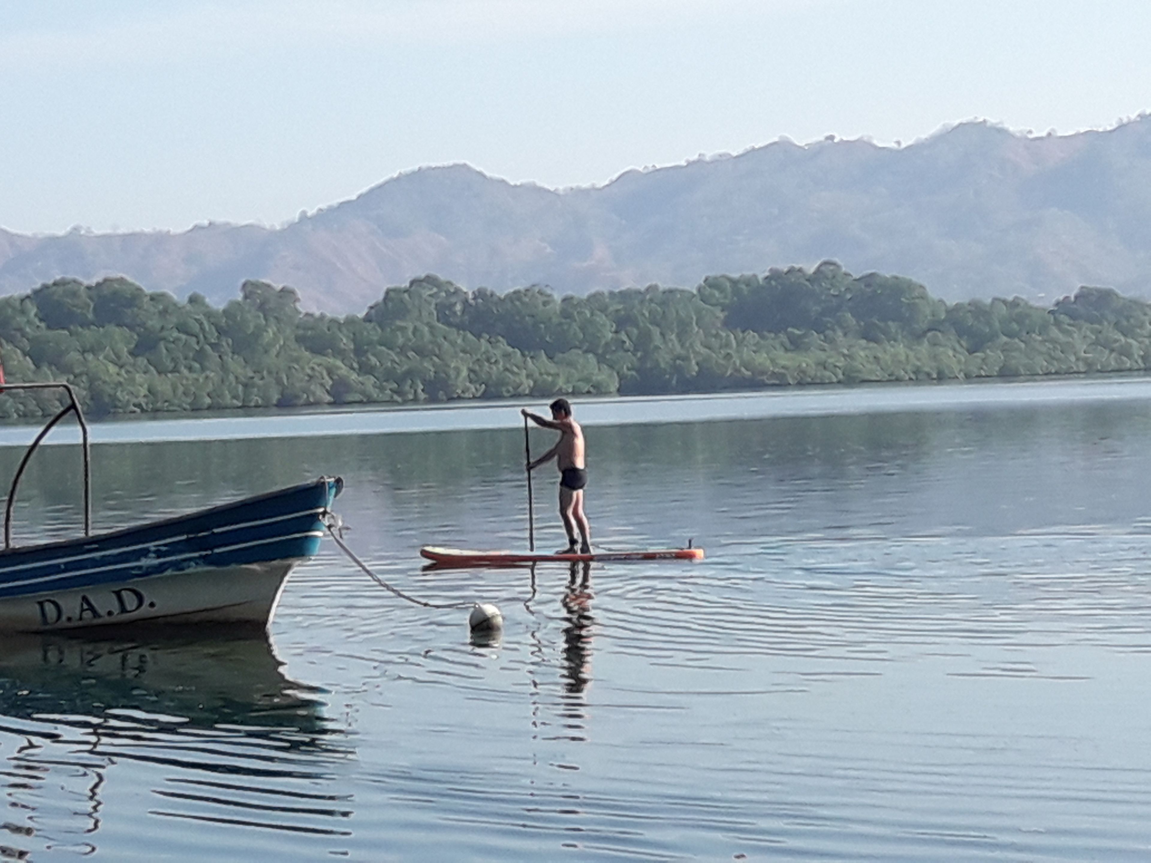 Una persona de pie en una tabla de remo en un lago tranquilo, con un barco llamado "D.A.D." al frente y montañas en el fondo.