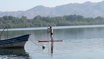 Una persona de pie en una tabla de remo en un lago tranquilo, con un barco llamado "D.A.D." al frente y montañas en el fondo.
