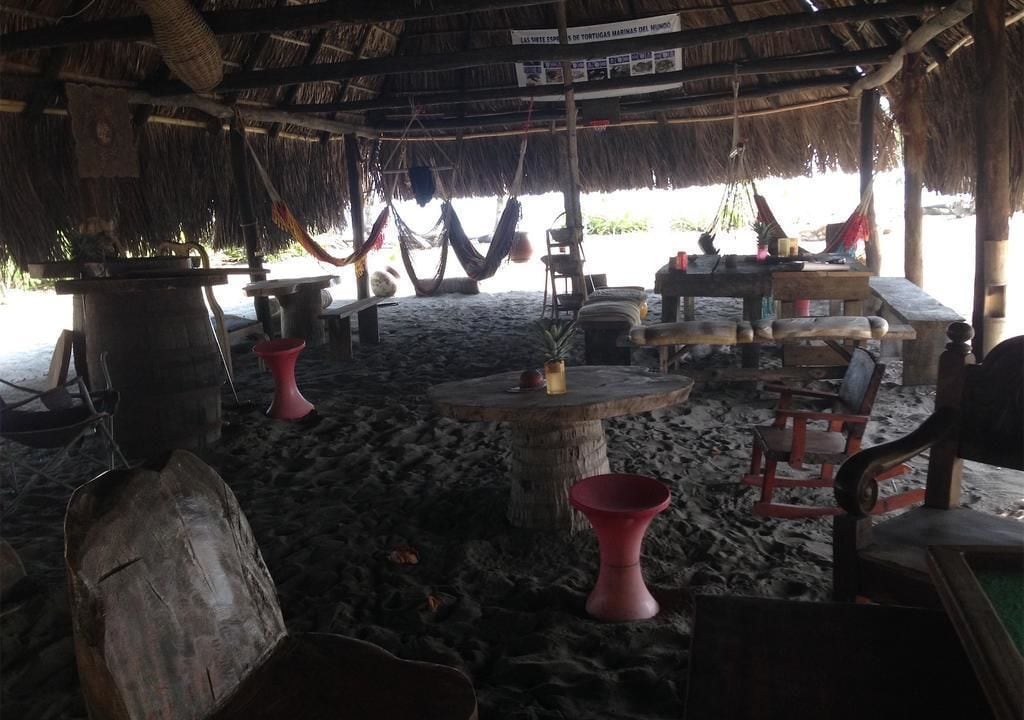 Una cabaña en la playa con techo de paja, muebles de madera y una olla grande sobre la arena.