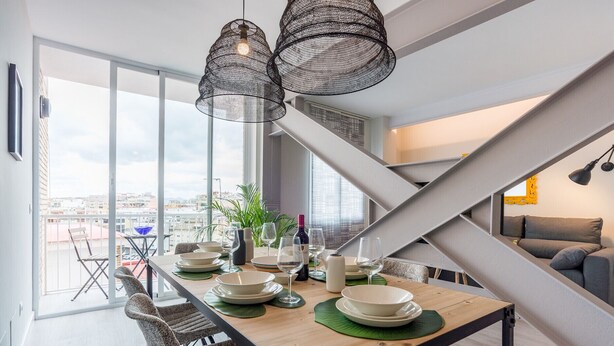 Un coin repas avec une table en bois dressée pour quatre personnes, entourée de chaises en osier. On aperçoit deux lampes suspendues, une plante en pot et une bouteille de vin sur la table. Un escalier avec une rampe en métal est visible à l’arrière-plan.