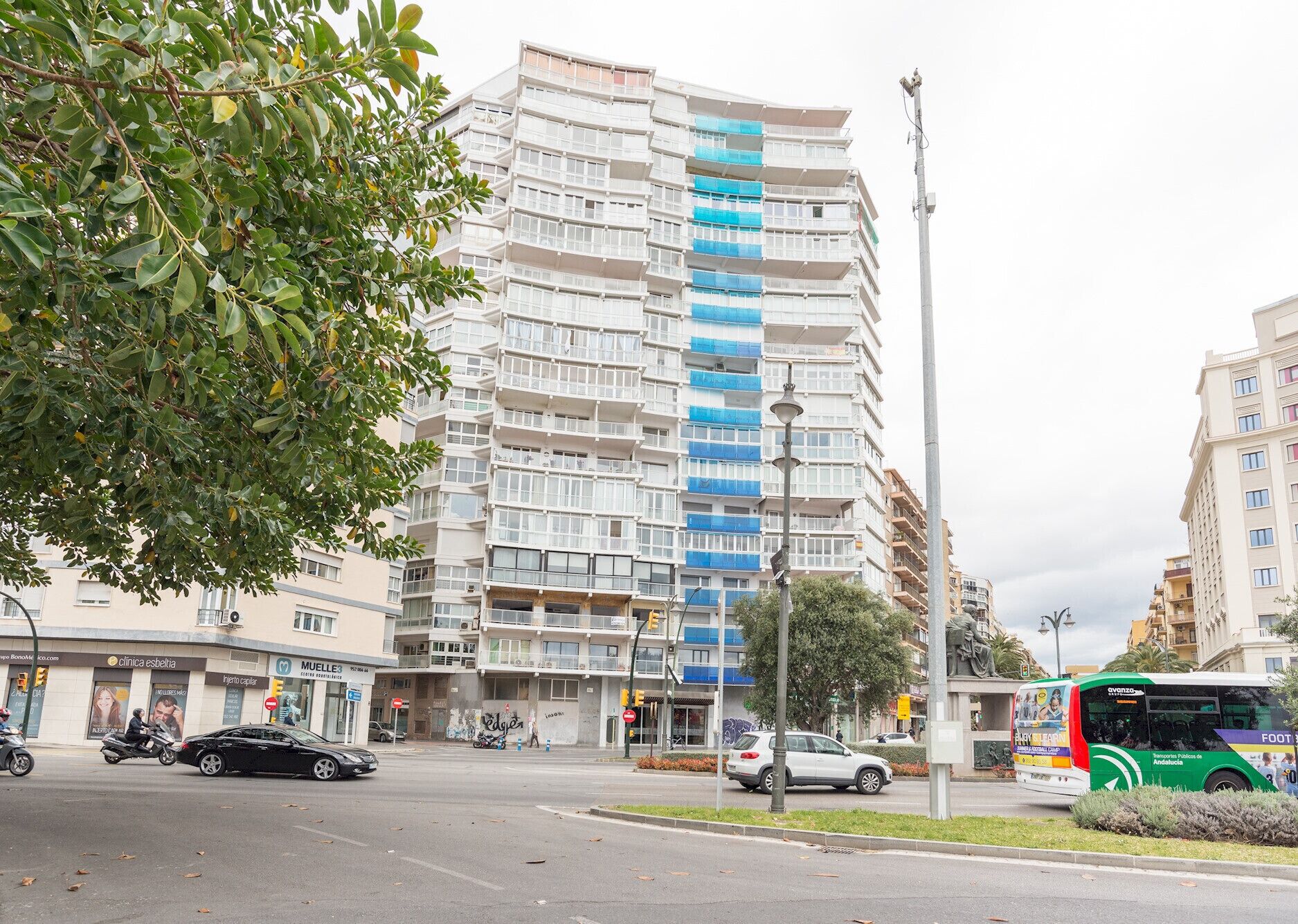 Un edificio de varios pisos con balcones, una calle con autos y un autobús, y un árbol en primer plano.