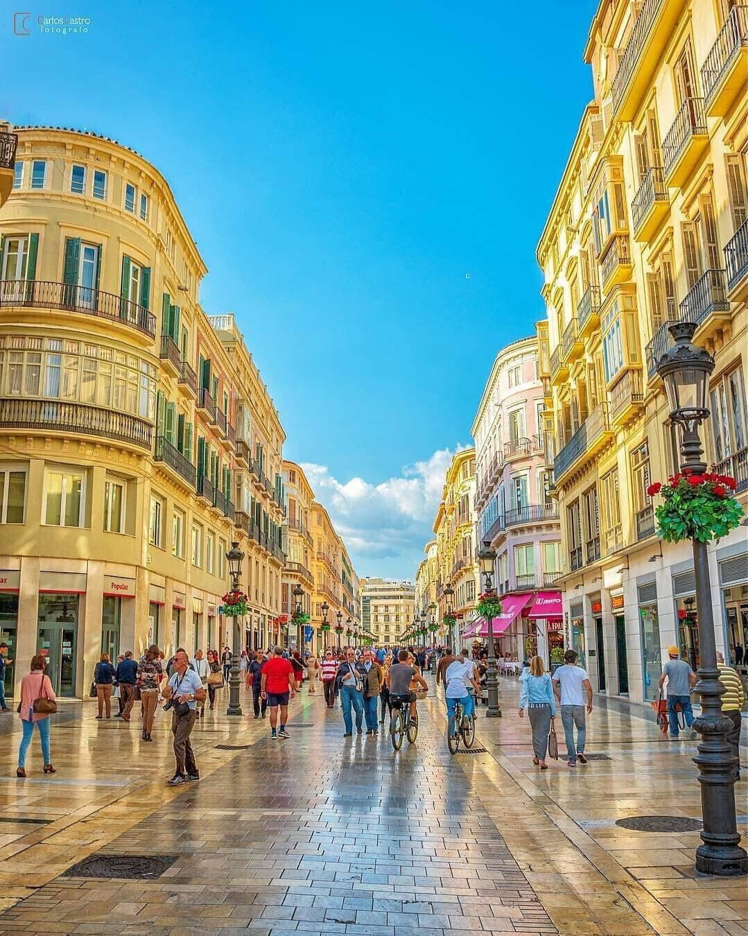 A wide pedestrian street lined with historic buildings, featuring a central open space with people walking and cycling.