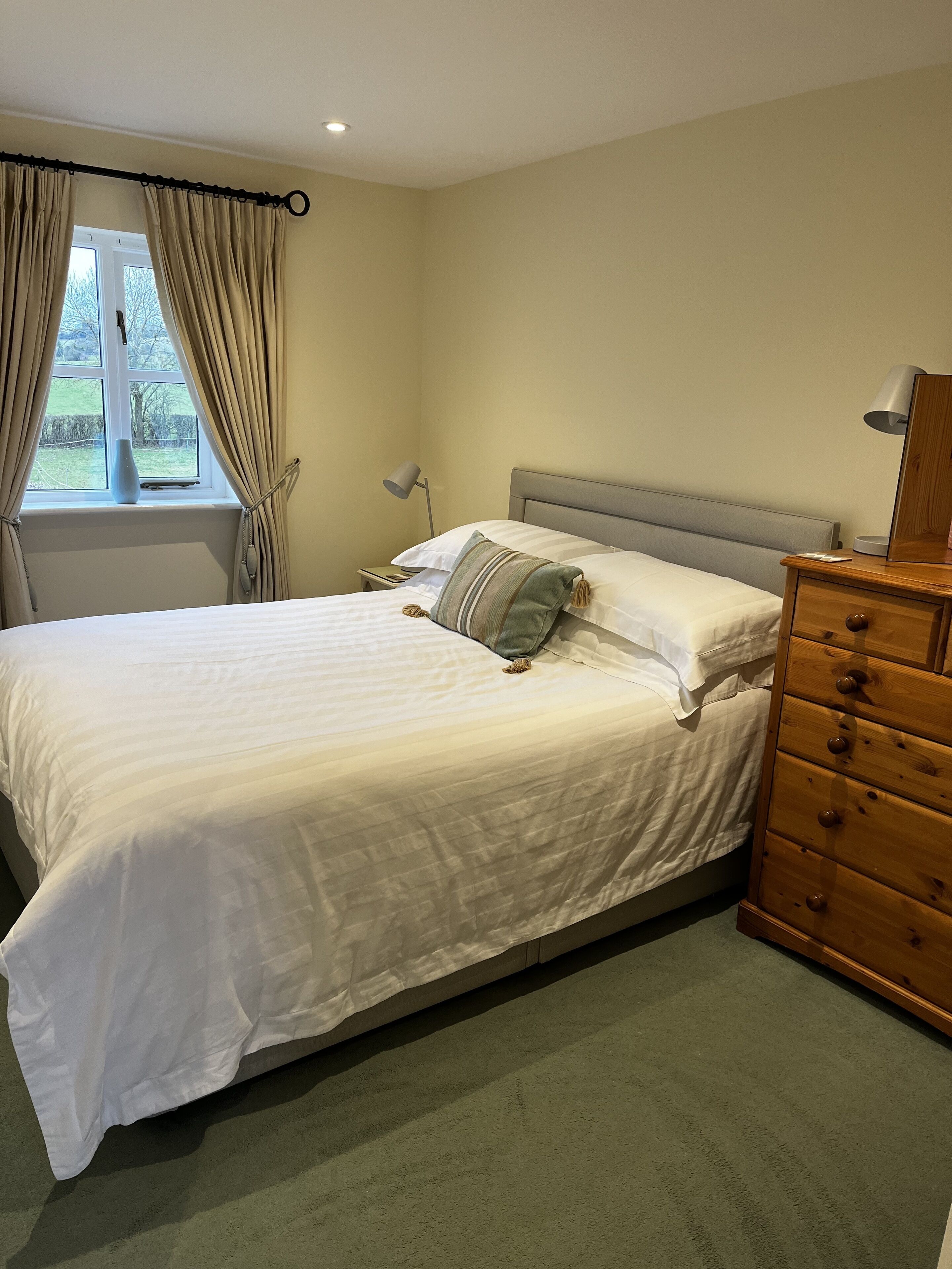 A neatly made bed with white linens, a wooden dresser with drawers, a bedside lamp, and a window with curtains.