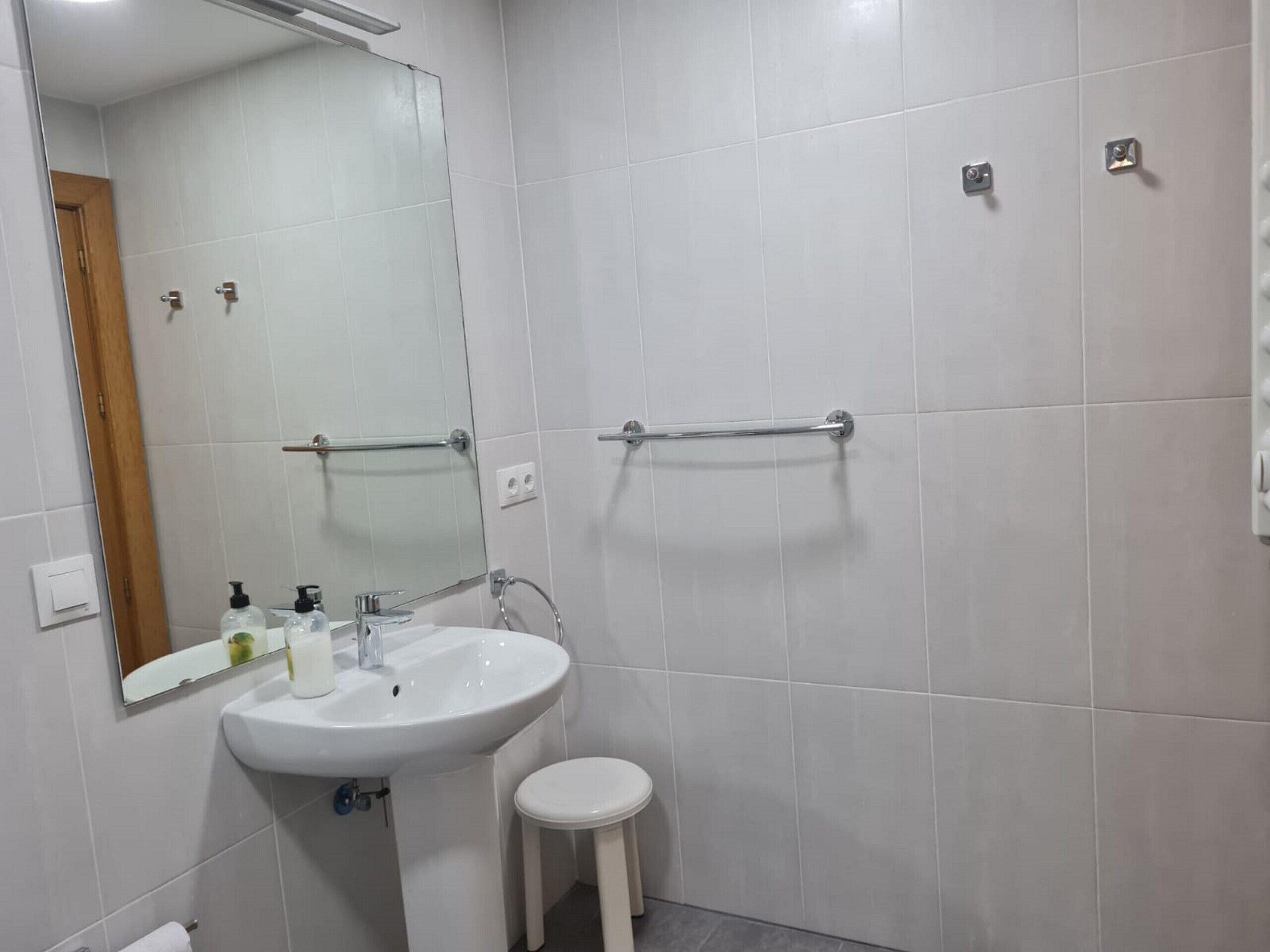 Een badkamer met een witte wastafel, spiegel en toilet. Er zijn handgrepen aan de muur en een klein krukje naast het toilet.