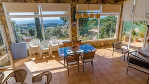 A sunlit patio with a table and chairs, large windows offering a view of the sea and greenery, and stone walls.