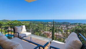 A balcony with a glass table and chairs, overlooking a coastal landscape with a clear view of the sea and greenery.