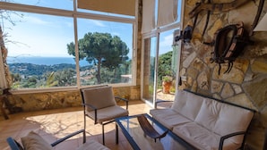 A sunlit room with a stone wall, a sofa, two chairs, and a glass table. A large window offers a view of a tree and distant landscape.
