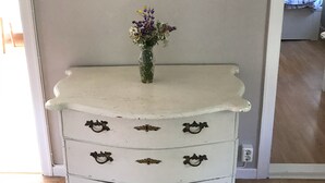 Une commode blanche ornée de poignées métalliques décoratives, une horloge murale en bois au-dessus, et un vase avec des fleurs posé sur le dessus.
