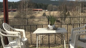 Une terrasse en bois avec une table et deux chaises, d’où l’on peut admirer un paysage rural avec des arbres et un bâtiment rouge situé au loin.
