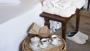 A neatly made bed with white linens, a bedside table with a lamp, a small table with towels and a book, and a wicker basket with tea set.