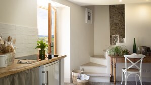 A kitchen with a wooden countertop, a dishwasher, a window with a view, a stone wall, and a dining area with a chair and table.