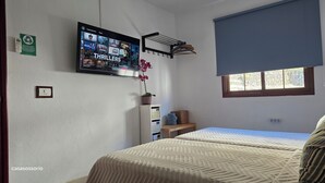 Een slaapkamer met twee bedden, een aan de muur bevestigde televisie waarop een film wordt afgespeeld, een raam met uitzicht en een plank met spullen.