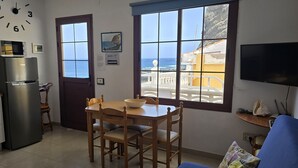 Een compacte keuken met een koelkast, magnetron en een eettafel met stoelen. Een groot raam biedt uitzicht op de zee en een balkon.